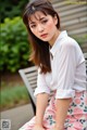 A woman sitting on a bench wearing a white shirt and floral skirt.