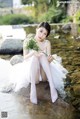 A woman in a white dress sitting on a rock by a river.