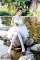 A woman in a white dress sitting on a rock by the water.